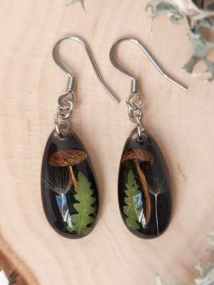 Close-up top view of two black resin earrings with mushrooms, fern leaves, and dandelion seeds arranged on a wood slice.