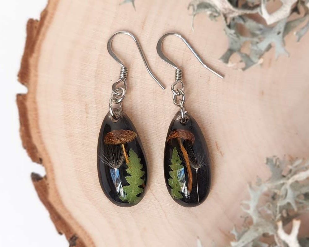 Close-up top view of two black resin earrings with mushrooms, fern leaves, and dandelion seeds arranged on a wood slice.