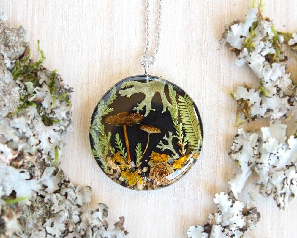 Close-up of a black resin pendant with real yellow lichen, ferns, and mushrooms on a light wood background surrounded by natural textures.