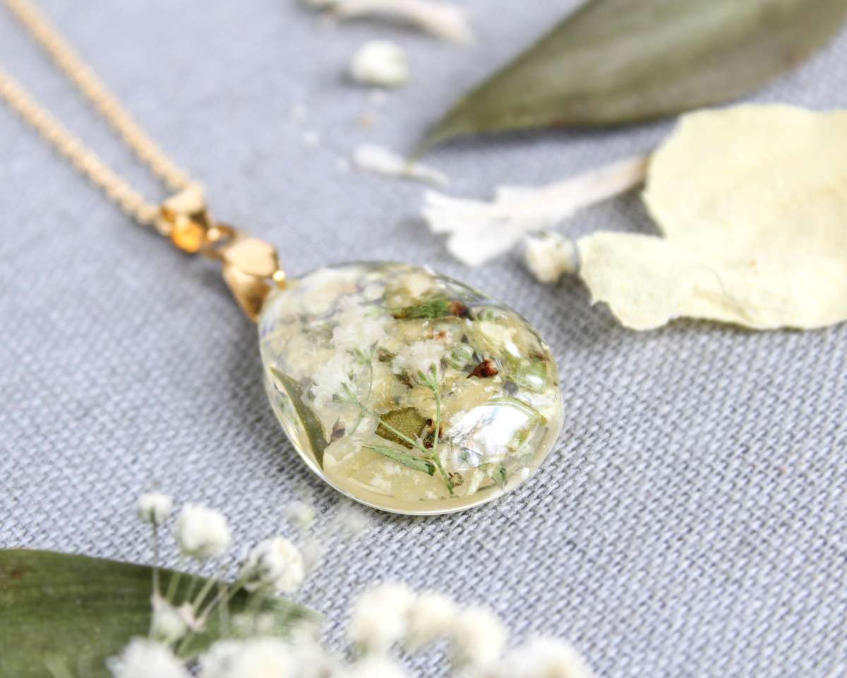 Close-up of a resin necklace lying on fabric, featuring preserved white wedding petals and greenery inside a teardrop pendant.