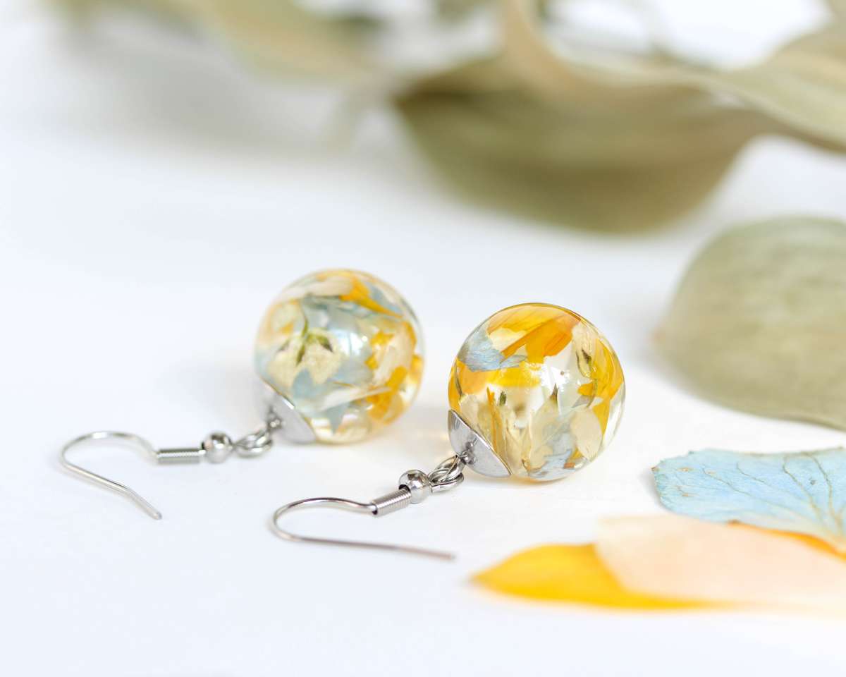 Pair of round resin earrings made with yellow, blue, and cream dried flower petals, displayed on a white surface with scattered petals