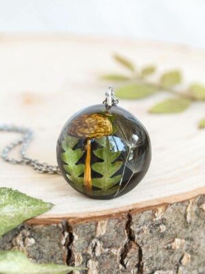 Artisan pendant with encapsulated mushroom and fern, placed vertically on a smooth wood slice with delicate leaf accents