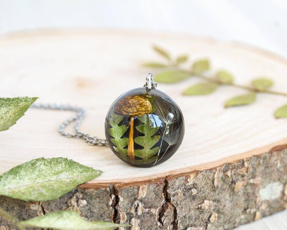 Artisan pendant with encapsulated mushroom and fern, placed vertically on a smooth wood slice with delicate leaf accents