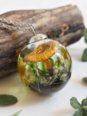 Close-up of a round resin pendant resting on wood, encasing a golden mushroom, yellow mimosa blossoms, and green botanical textures.
