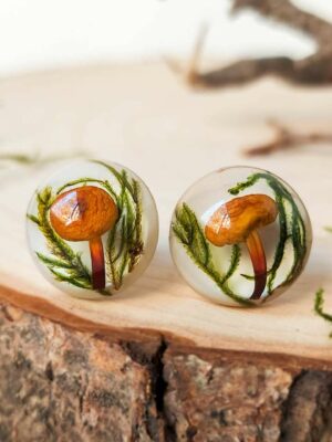 Pair of round resin stud earrings with real orange mushrooms and moss, displayed on a wood slice.