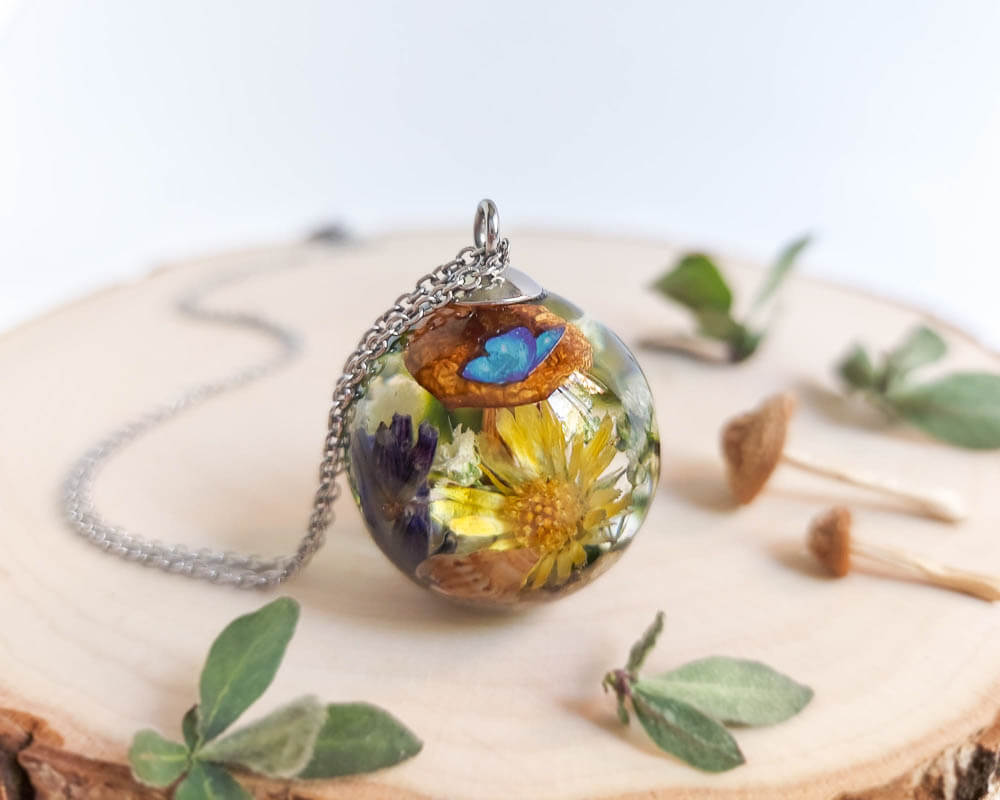Pendant on wooden surface showcasing a vivid arrangement of yellow petals, mushroom, and butterfly wing inside resin.