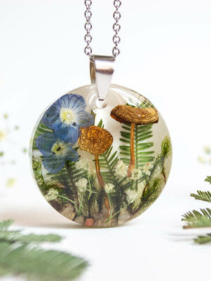 Round botanical resin necklace with preserved blue flowers, two mushrooms, ferns, and white wildflowers inside, hanging on a stainless steel chain