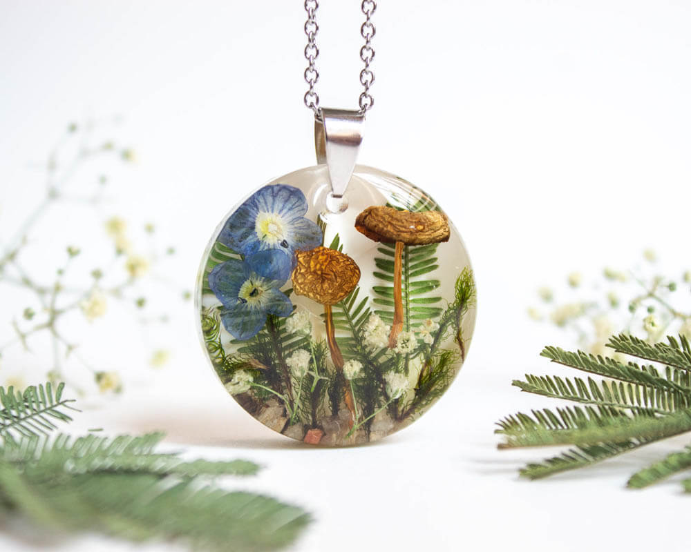 Round botanical resin necklace with preserved blue flowers, two mushrooms, ferns, and white wildflowers inside, hanging on a stainless steel chain