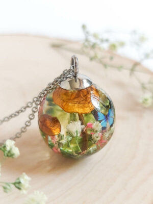 Handmade spherical pendant showcasing a miniature natural landscape with mushrooms, flowers, and a butterfly, displayed on a wooden slice.