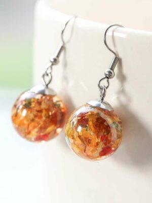 Spherical resin earrings with red, yellow, and orange preserved wedding petals, hanging from the rim of a white ceramic cup