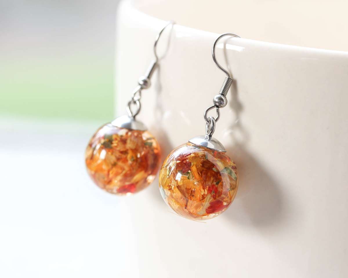 Spherical resin earrings with red, yellow, and orange preserved wedding petals, hanging from the rim of a white ceramic cup