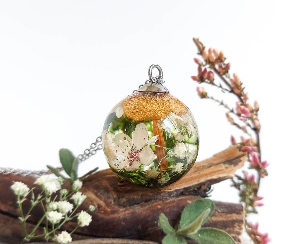 Resin necklace with cherry blossom, moss, and mushroom displayed on wood and surrounded by small flowers