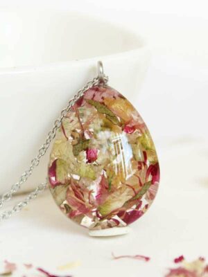 Teardrop-shaped resin pendant necklace with pressed flowers in pink, ivory, and green, photographed in front of a white bowl.